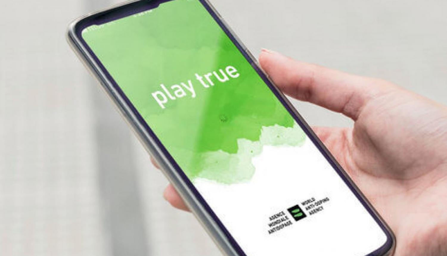 Cilvēka plauksta, kas rokās tur viedtālruni, kura ekrānā ir attēlots attēls ar tekstu "Play True"